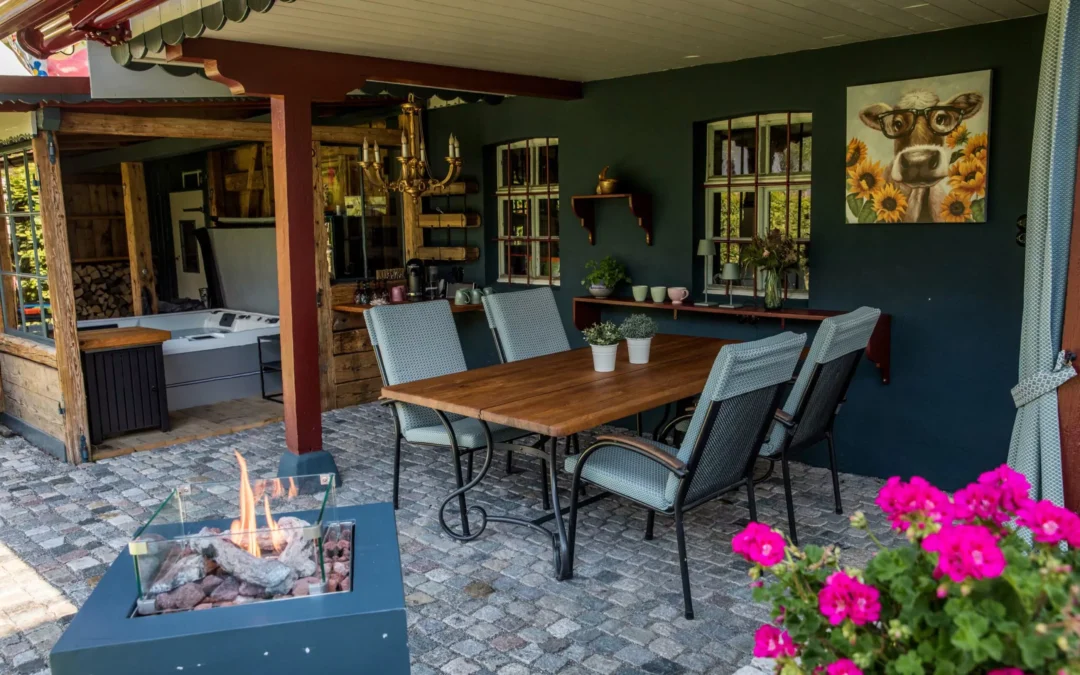 Holztisch mit Stühlen auf einer Terrasse, umgeben von Pflanzen und einer Feuerstelle.