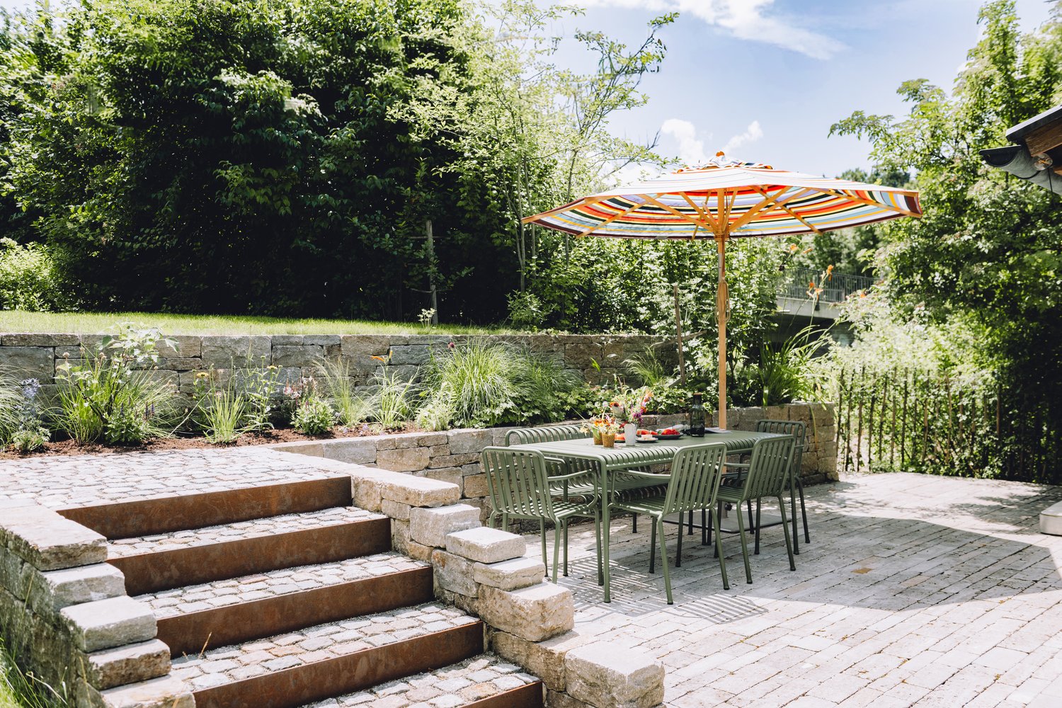 Ein farbenfroher Sonnenschirm schützt den Gartentisch auf der gepflasterten Terrasse zwischen Natursteinmauern und üppigem Grün von Hübner.