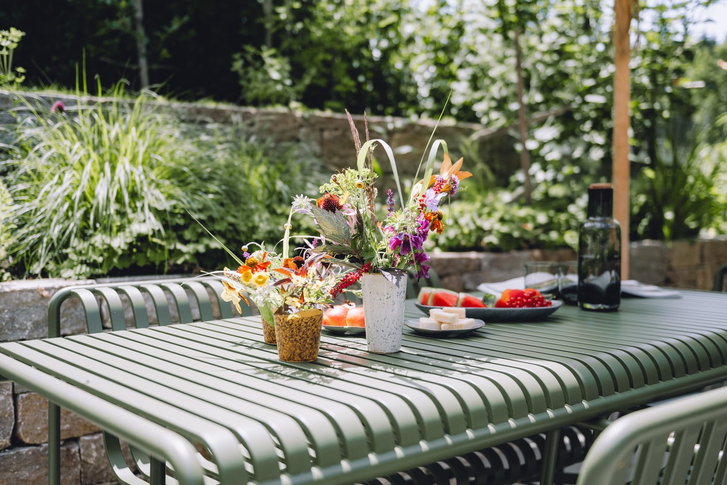 Im Garten steht ein grün gedeckter Esstisch mit Blumenarrangements, Obst, Wasser und Geschirr für ein entspanntes Sommeressen im Freien unter Bäumen.