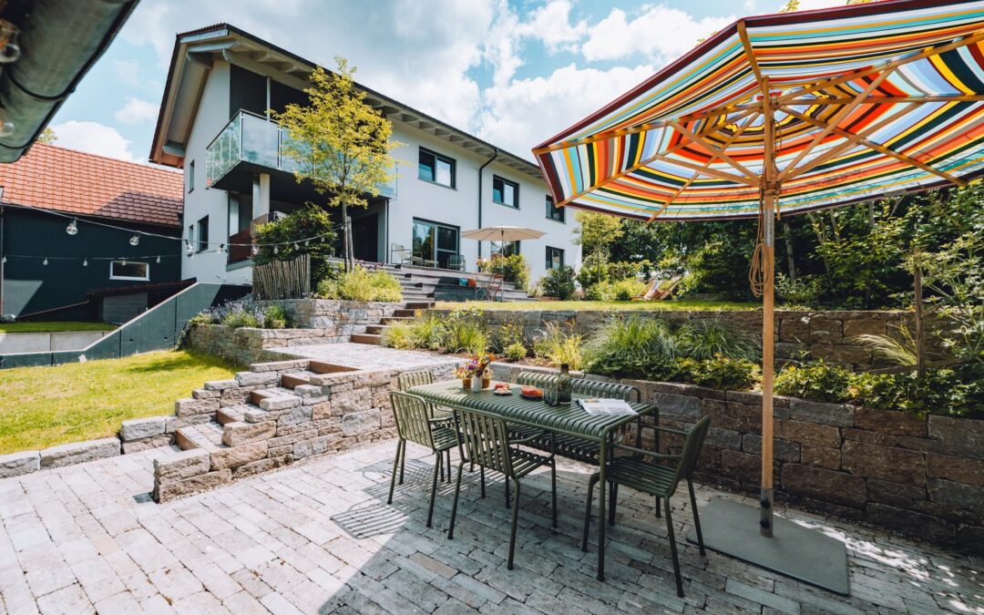 Auf der Terrasse stehen ein bunter Sonnenschirm, ein Tisch mit Stühlen und ein modernes Haus mit Balkon, umgeben von Stufen und Bepflanzung.