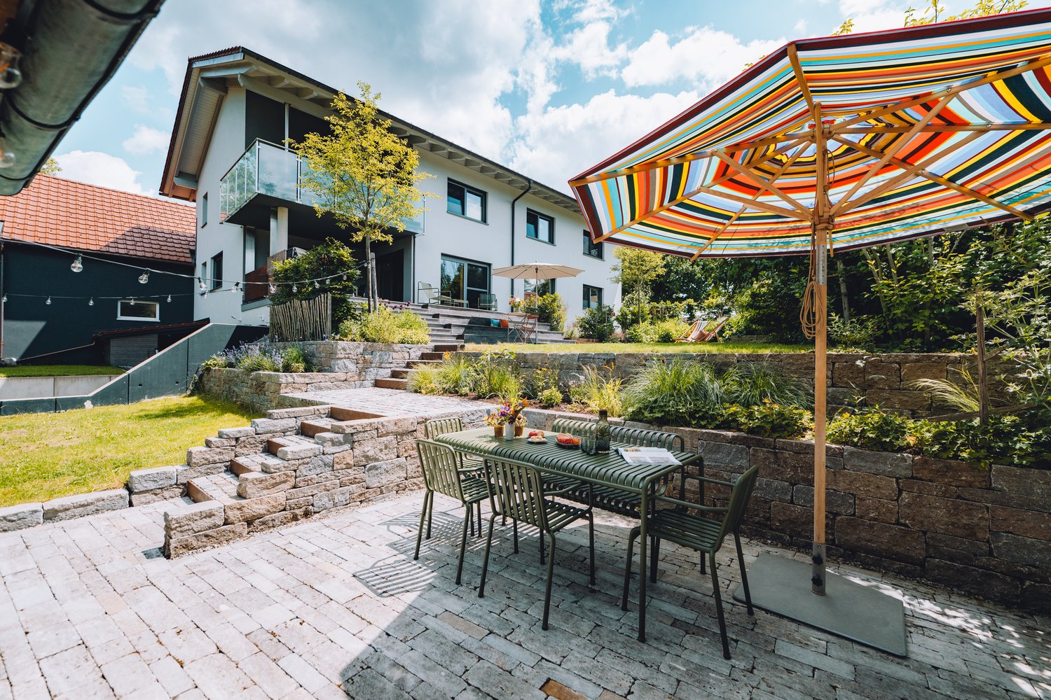 Auf der Terrasse stehen ein bunter Sonnenschirm, ein Tisch mit Stühlen und ein modernes Haus mit Balkon, umgeben von Stufen und Bepflanzung.
