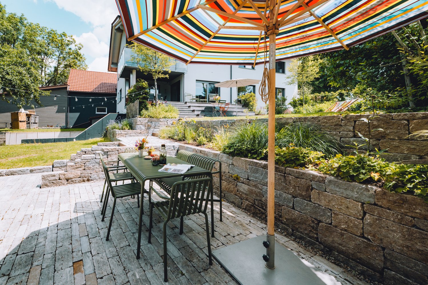Eine moderne Gartenterrasse mit grünem Tisch, Streifen-Sonnenschirm, Natursteinmauern und bepflanzten Ebenen vor einem Wohnhaus.