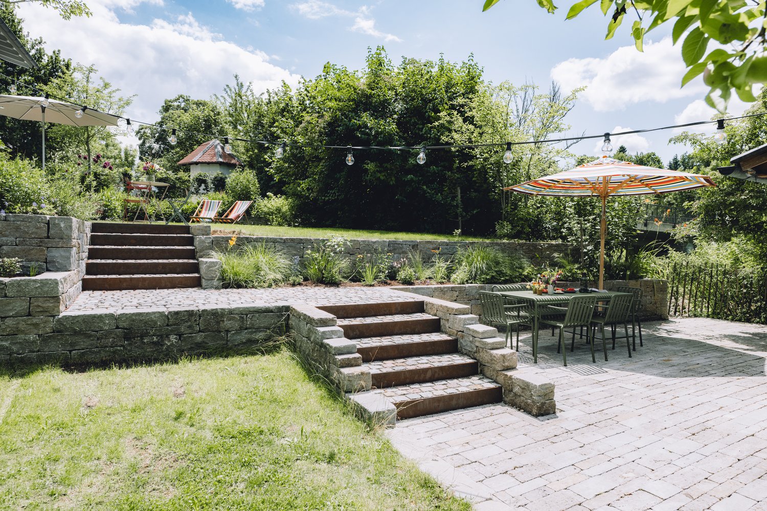 Eine liebevoll gestaltete Gartenterrasse mit Natursteinmauern, Stufen, Pflasterfläche, Sitzgruppe und bunten Sonnenschirm liegt unter grünem Blätterdach.