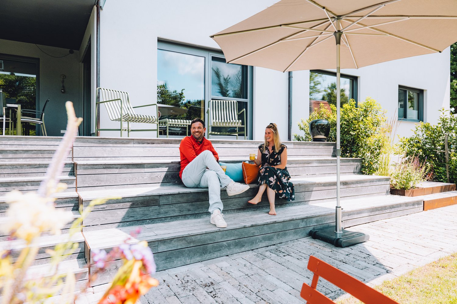 Ein Paar sitzt entspannt auf einer modernen Holzterrasse mit großem Sonnenschirm, Getränken und Kissen vor einem hellen Haus mit Garten und Pflanzbeeten.