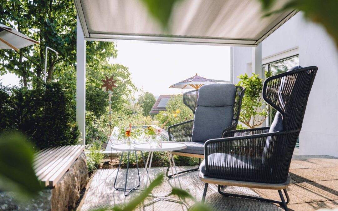 Auf der geschützten Terrasse stehen moderne Lounge-Sessel mit Beistelltisch, umgeben von Grün, Sonnenschirm und Blick in den Garten.