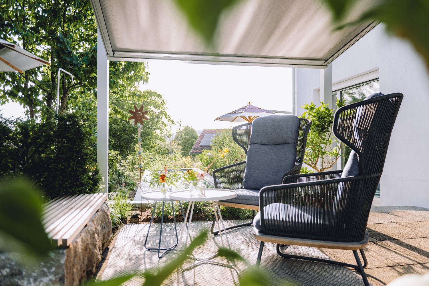Auf der geschützten Terrasse stehen moderne Lounge-Sessel mit Beistelltisch, umgeben von Grün, Sonnenschirm und Blick in den Garten.