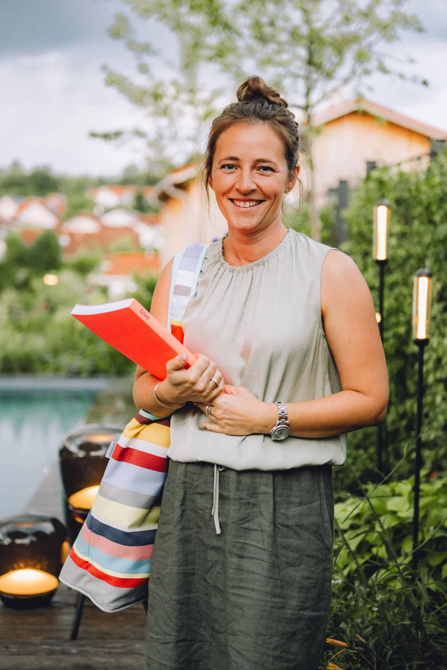 Frau mit rotem Buch im Garten, Hübner Traumgärten und Abendstimmung Eine lächelnde Frau hält ein rotes Buch im Garten, während im Hintergrund Häuser, Bäume und sanfte Lichter entlang des Weges zu sehen sind.