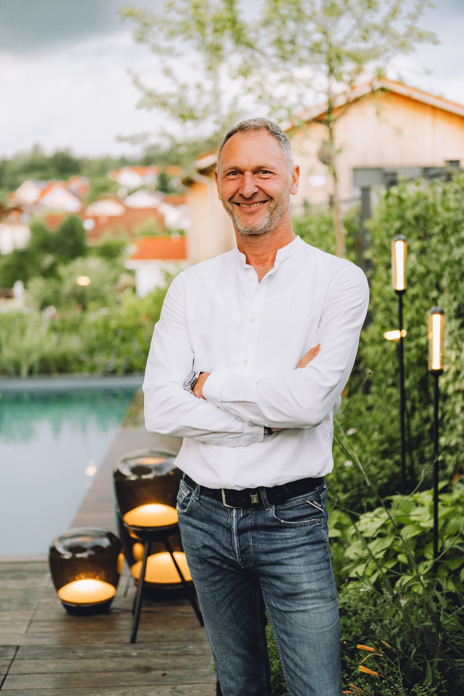 Ein lächelnder Mann steht mit verschränkten Armen in einem modernen Garten mit Pool, Beleuchtung und üppiger Bepflanzung vor dem Haus.