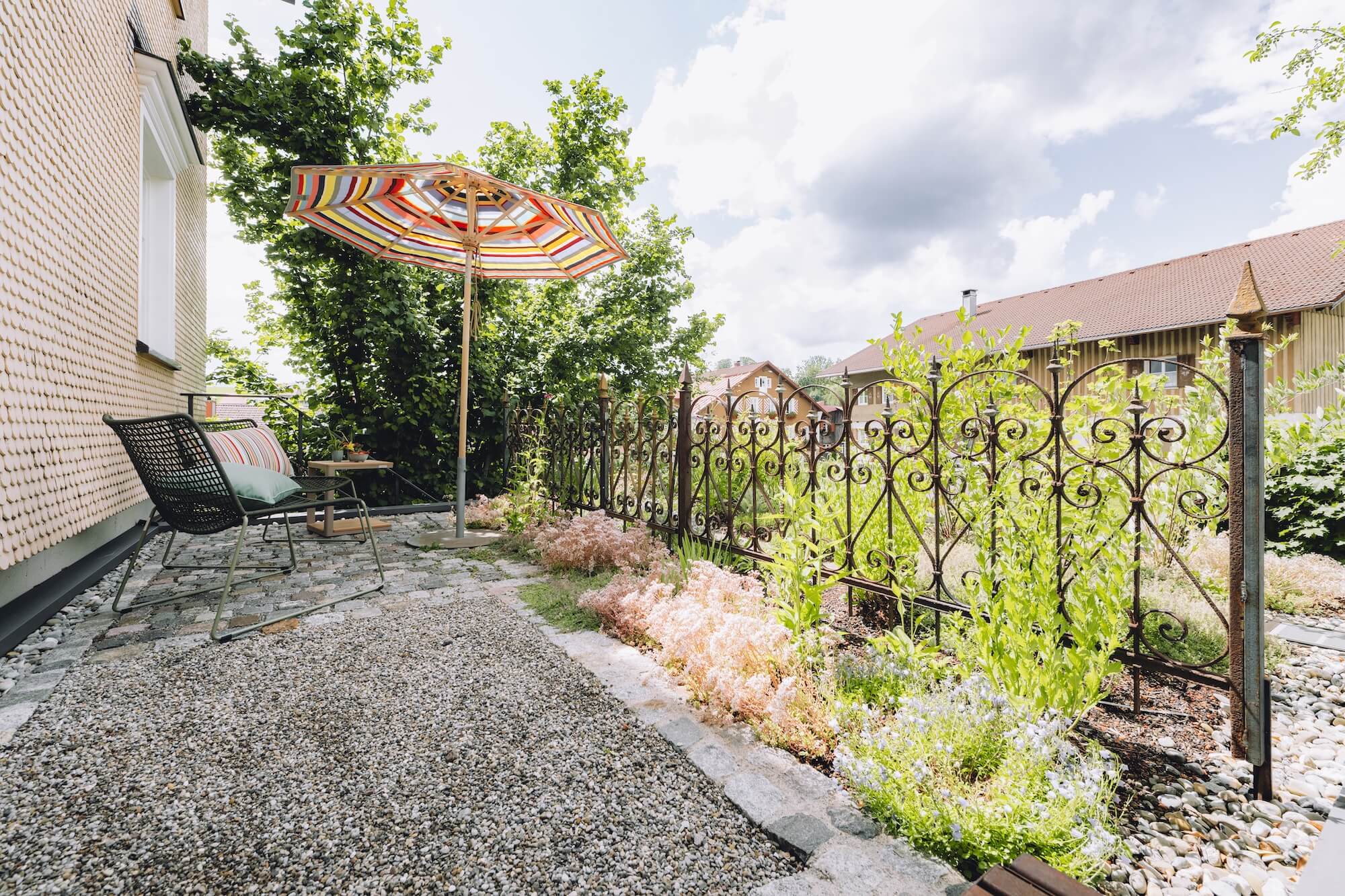 Kleiner Dachgarten mit Streifen-Sonnenschirm, Naturstein und Schmiedeeisen Ein Paar sitzt entspannt auf einer modernen Holzterrasse mit großem Sonnenschirm, Getränken und Kissen vor einem hellen Haus mit Garten und Pflanzbeeten.