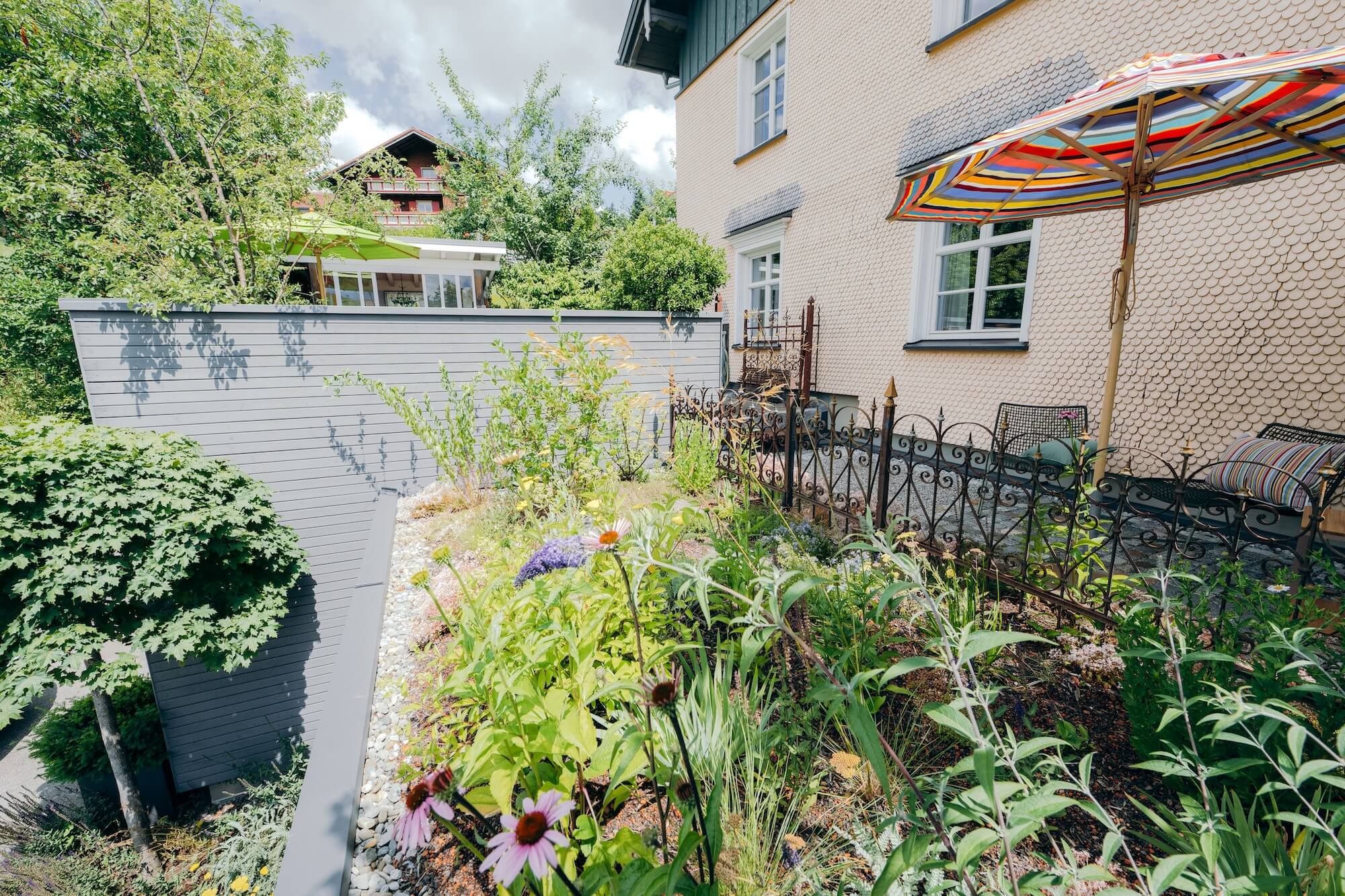 Kleiner Dachgarten mit Streifenschirm und Stauden in Stiefenhofen Unter einem bunten Streifenschirm steht auf der Garten-Terrasse ein grüner Esstisch mit Stühlen, umgeben von Steinmauer, Stauden und viel Grün.