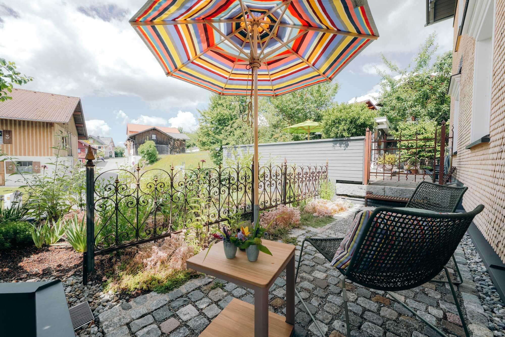Kleiner Dachgarten mit bunt gestreiftem Sonnenschirm und Natursteinpflaster Eine moderne Gartenterrasse mit grünem Tisch, Streifen-Sonnenschirm, Natursteinmauern und bepflanzten Ebenen vor einem Wohnhaus.