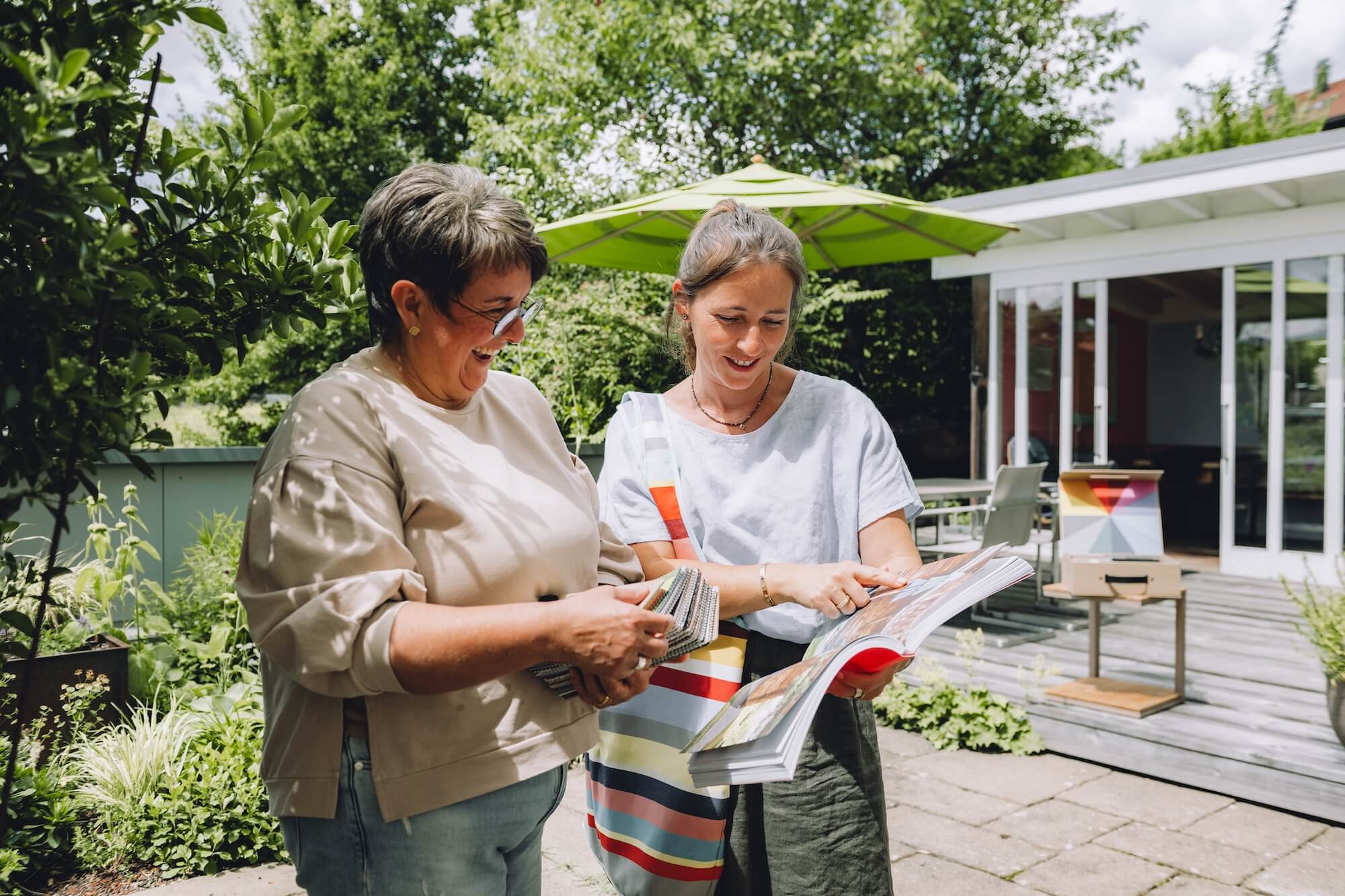 Beratung im Dachgarten von Hübner in Stiefenhofen, Westallgäu Im Garten steht ein grün gedeckter Esstisch mit Blumenarrangements, Obst, Wasser und Geschirr für ein entspanntes Sommeressen im Freien unter Bäumen.