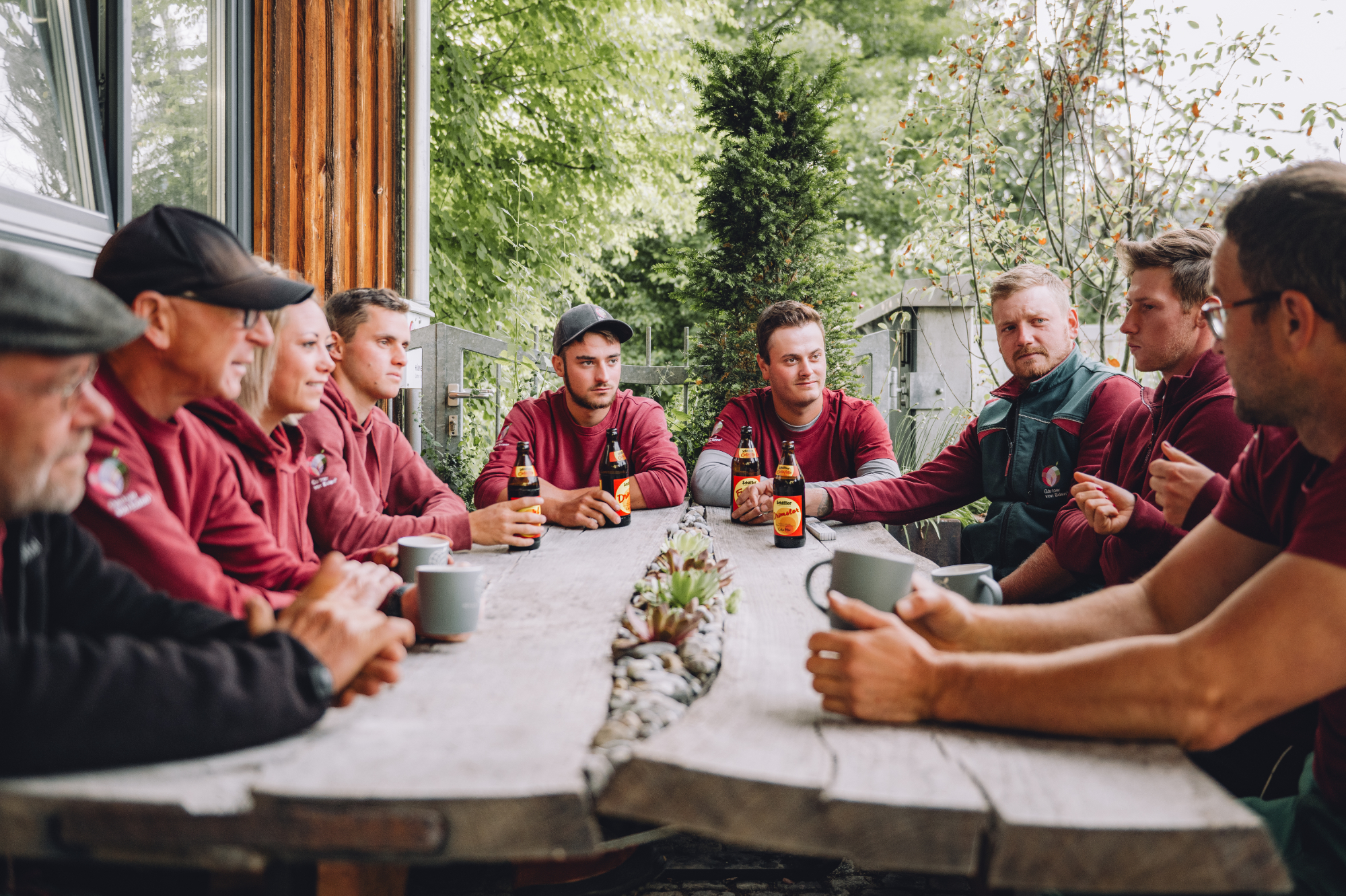 Gemeinsame Pause des Hübner-Teams am rustikalen Gartentisch Zwei Gärtner stehen und sitzen am Rand eines Pools in einem Garten.